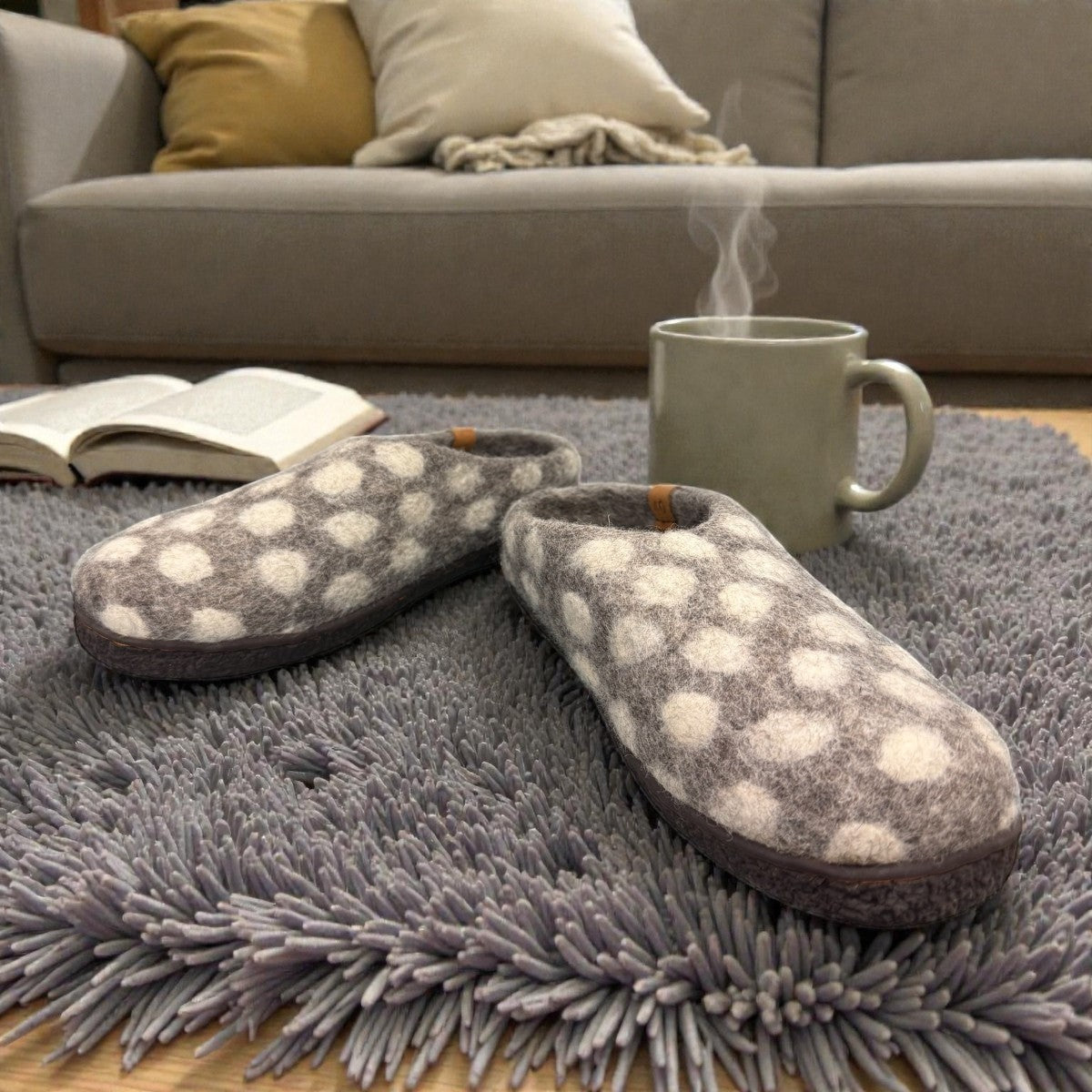 Lhasa grey wool mule slippers with white polka dots on a grey rug next to a steaming mug
