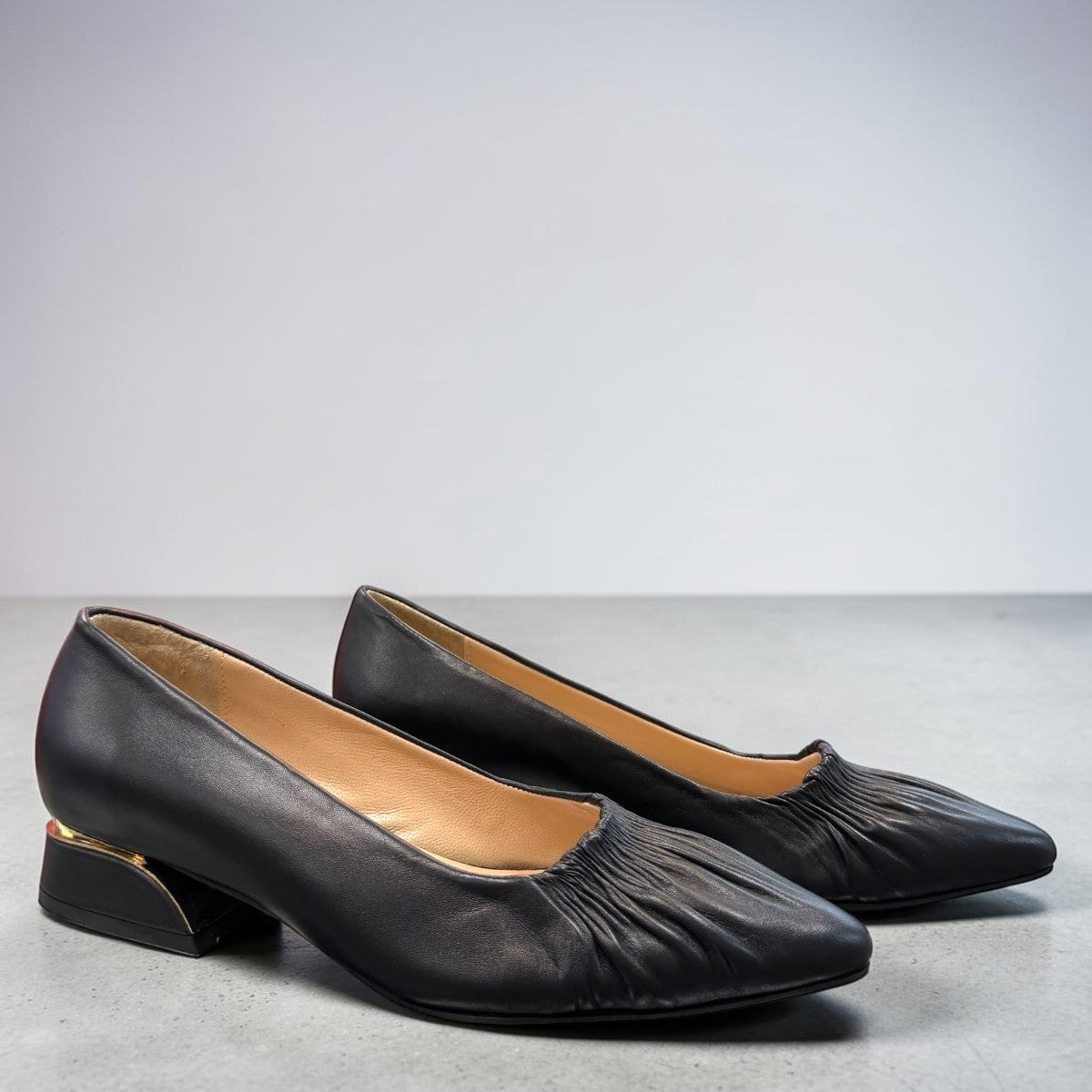 LouJo Betty black leather pump with a pointed toe, pleated detail, and a low sculpted metallic heel.