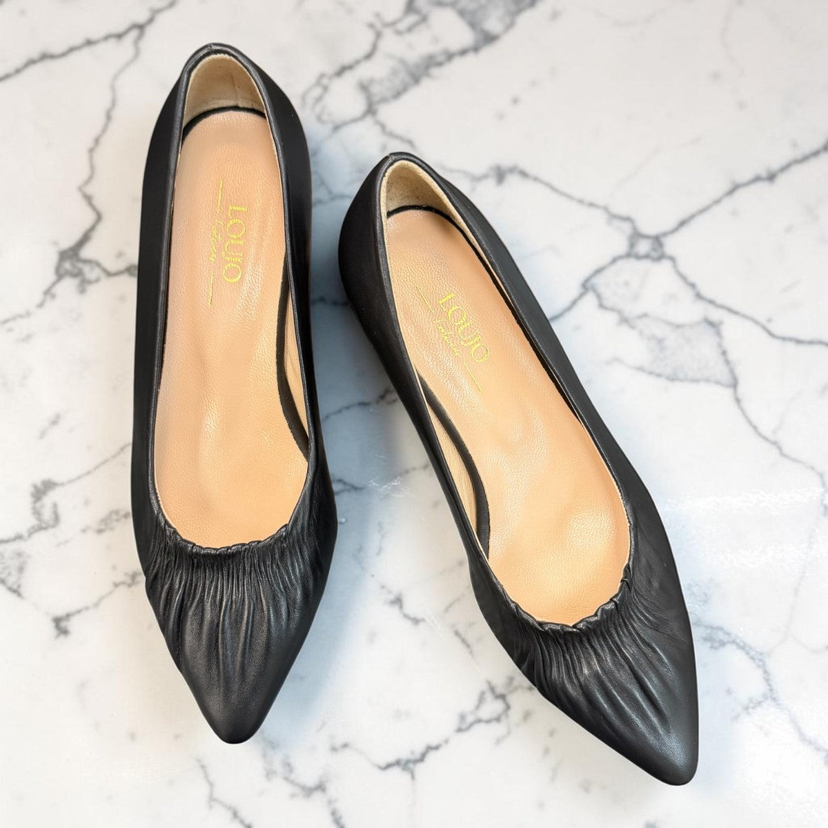 LouJo Betty black leather pump with a pointed toe, pleated detail, and a low sculpted metallic heel.