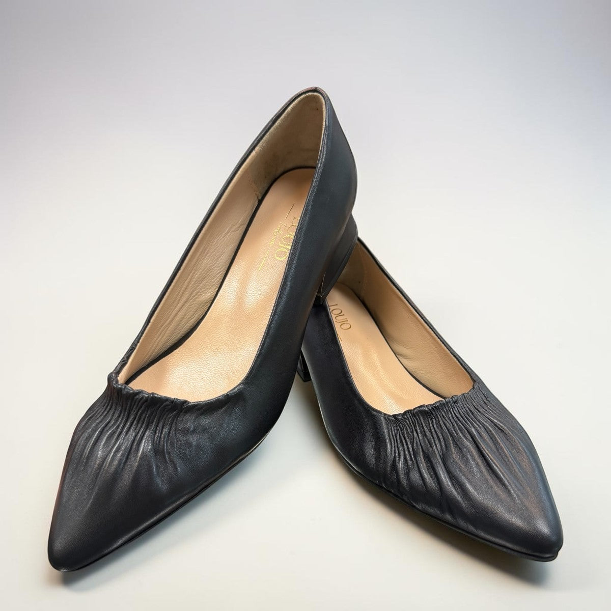 LouJo Betty black leather pump with a pointed toe, pleated detail, and a low sculpted metallic heel.