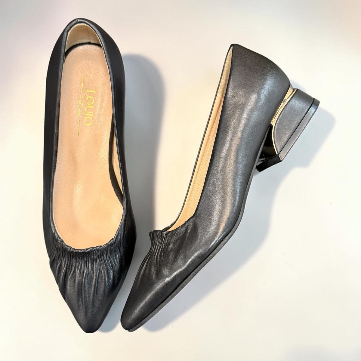 LouJo Betty black leather pump with a pointed toe, pleated detail, and a low sculpted metallic heel.